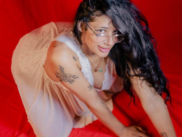 BonnyEbonny de quatro, usando óculos, um vestido branco e mostrando tatuagens nos braços. Ela tem cabelo escuro ondulado, um sorriso fofo e um colar. Suas nádegas estão visíveis..