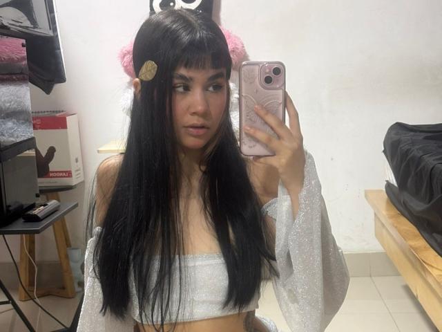 IsabellaMarynn with long black hair, silver off-shoulder top, holding pink phone, looking at mirror, pink hairclip, small tits, slim, pale skin, bedroom, wooden bed, black bag.