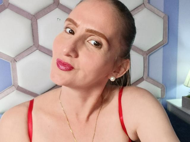 RubiAnderson has shiny pink lipstick, a gold necklace, and red straps, with her brown hair pulled back, and is making a playful, pouty face with her eyebrows raised.