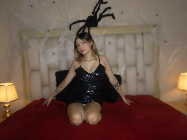 TaniaRico with small tits, black shiny dress, and tattoos, kneeling on red bed, black spider headband, flirty expression, lamps on both sides.