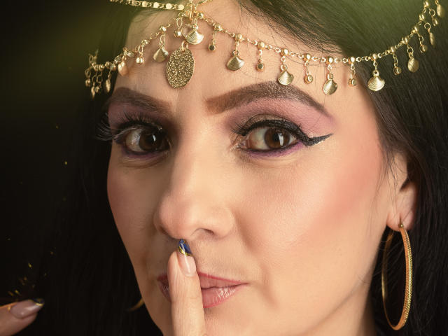 JakyKidman's face with dark hair, wearing a gold headpiece with dangling coins, has purple eyeshadow, blue nail polish, and a finger covering her nose. She's sporting gold hoop earrings.