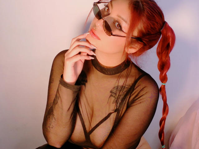 Ameliyes with red ponytail, wearing black sheer top, showing her tits, hand near mouth, brown glasses, pink lipstick, white wall, and a nose ring in a sexy, confident pose.