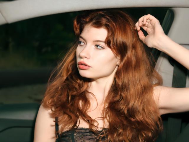 LoliyPop with long, wavy red hair, pale skin, and blue eyes, wearing a black lace top, sits in a car, hand in hair, looking to the side.