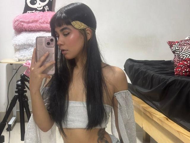 IsabellaMarynn, con lunghi capelli neri, indossa una maglietta argentata con una spalla scoperta, tiene in mano un telefono rosa, mostrando i suoi piccoli seni e un tatuaggio sul fianco, mentre guarda uno specchio..
