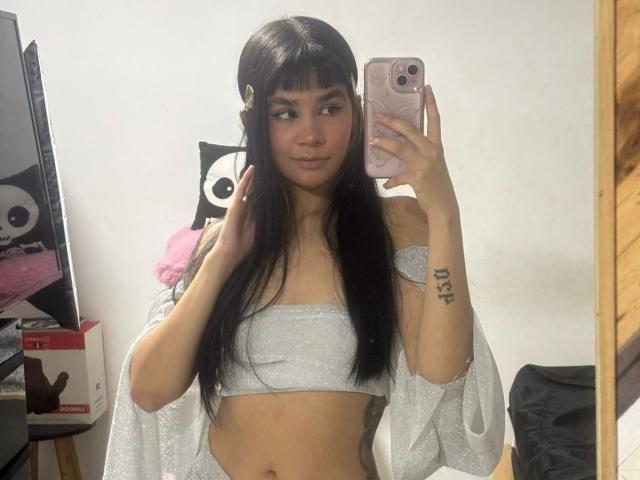 IsabellaMarynn stands in front of a mirror, holding a pink phone with a white and pink case. She has long black hair with bangs, light brown skin, and a tattoo on her right forearm. She wears a sparkly silver bandeau top and matching shawl with loose sleeves. Her right hand touches her hair. The background includes a white wall with a black and white cartoonish eye and a wooden shelf with a pink plush toy and a red box. The room has a wooden wall on the right.