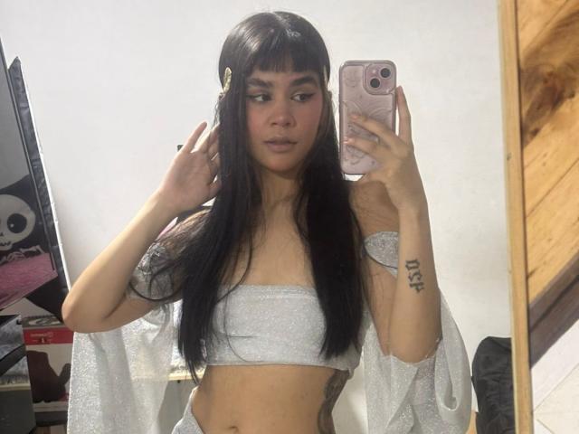 IsabellaMarynn, con lunghi capelli neri, indossa un top argento scintillante e pantaloni abbinati, sta scattando un selfie allo specchio, mostrando i suoi piccoli seni e un tatuaggio sulla mano destra..