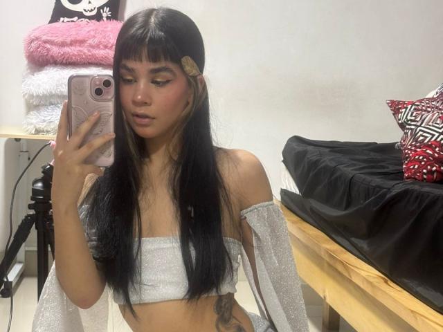 IsabellaMarynn, con lunghi capelli neri, indossa una top scintillante con una spalla scoperta, tiene in mano un telefono rosa, mostrando il suo seno piccolo e un tatuaggio, e sta scattando un selfie allo specchio in una camera da letto..