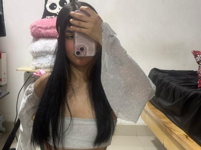 IsabellaMarynn takes a selfie in a mirror. She has long black hair, covering one eye with her pink phone. She wears a sparkly, off-shoulder, silver top with glittery sleeves. Her light skin contrasts with her dark hair. The background includes a stack of fluffy pink and white pillows on a shelf and a black bed with a red pillow. The image is casual and focused on IsabellaMarynn's upper body.