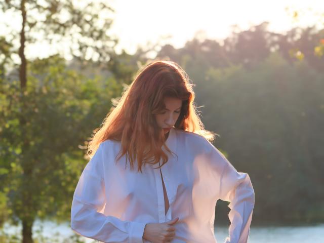 LoliyPop with red hair, wearing an open white shirt, exposing her tits, stands by a sunlit, green tree-filled area, hand in her hair, eyes closed, sunlight on her face.
