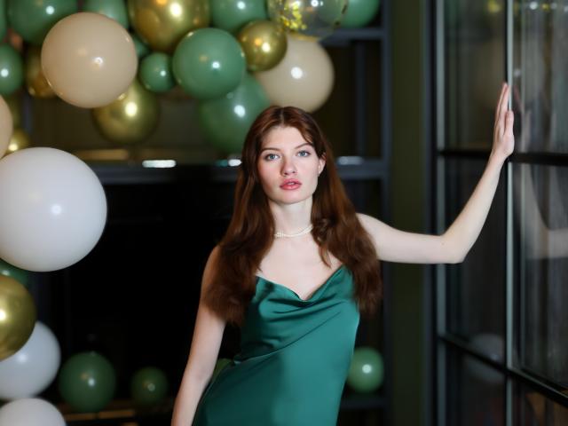 LoliyPop with long brown hair, green dress, and pearl necklace, stands by a window, touching the glass with her right hand, looking slightly to the side. Boobs are small.