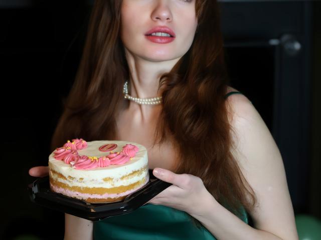LoliyPop with long red hair, blue eyes, and a pearl necklace holds a pink and white frosted cake with pink swirls on a black tray. She's wearing a green dress.