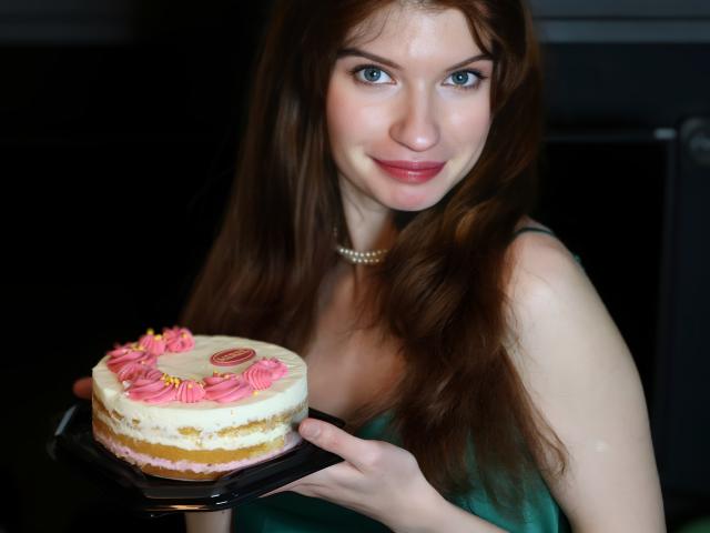 LoliyPop with long brown hair, pearl necklace, green dress, holding a pink and white layered cake with pink frosting, slightly parted pink lips, visible upper chest.