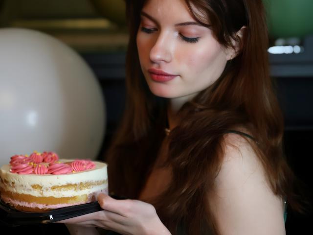 LoliyPop with brown hair, green dress, pearl necklace, holding a white cake with pink roses, standing in front of balloons. She's got pretty tits and a cute face.