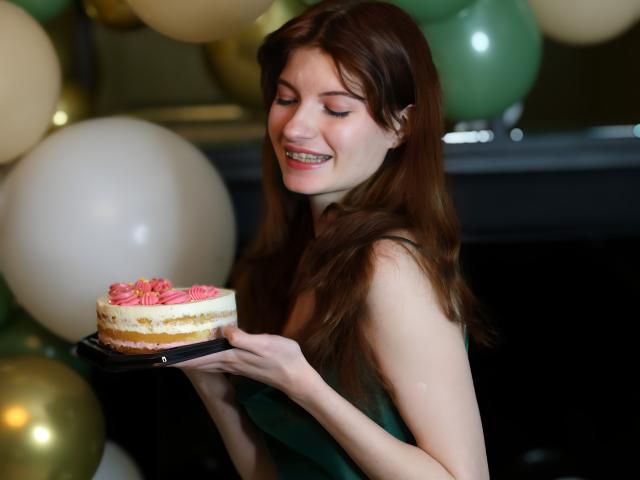 LoliyPop with long brown hair, holding a cake with pink swirled frosting, looking down, wearing a black top, in a dimly lit room with a white balloon.