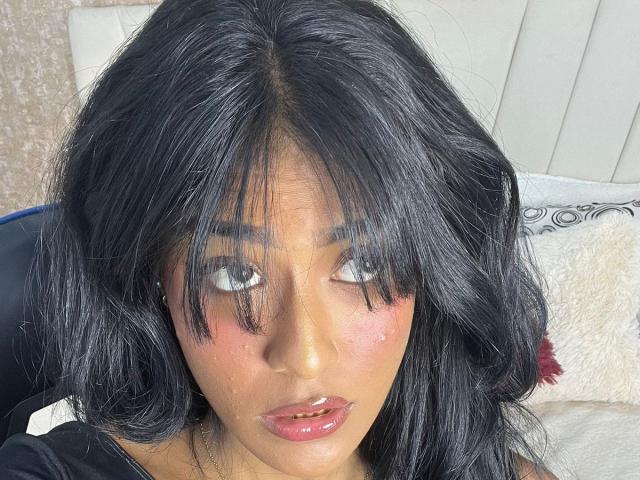 Babilett has dark black, wavy hair covering her eyes, flushed pink cheeks, and glossy pink lips. Her eyes are slightly upturned, looking off to the side.