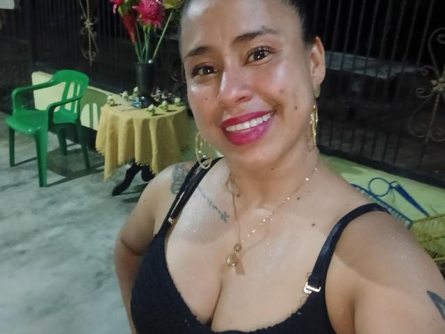 KamilaLovee with tan skin, black bra, showing her big boobs, pink lipstick, gold earrings, and necklace, smiling at the camera. She has tattoos on her chest.