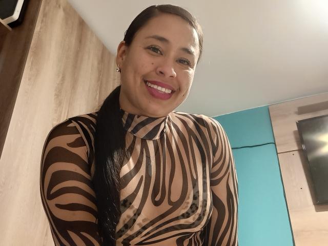 KamilaLovee with long black hair, smiling, wearing a zebra-patterned sheer top, showing her small, perky tits, pink lipstick, and earrings, in a well-lit room.