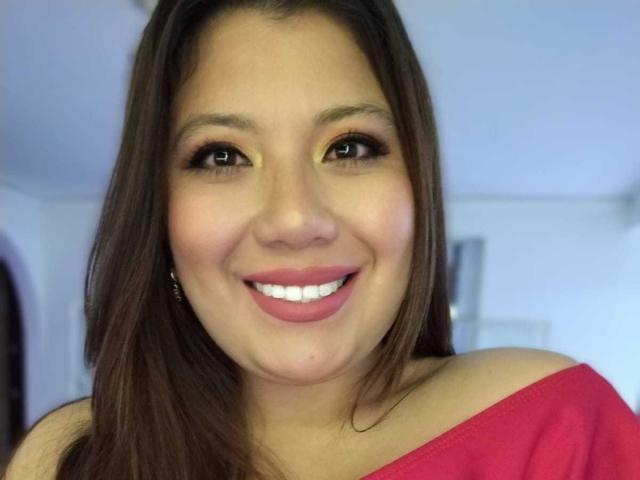 PaulaOwen with brown hair, yellow eye makeup, and pink lipstick, smiling, wearing a pink top, exposing her shoulder, has a cute, chubby face with a small nose.