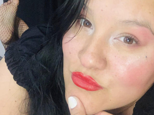 NuahElectra with red lipstick, white nails, black fishnet, and black hair, pouty face, brown eyes, slightly flushed skin, holding her chin, close-up, slight blush on cheeks.