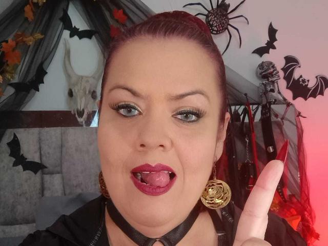 VanesaMoka has dark red lipstick, blue-green eyes, and is sticking out her tongue with a black choker, gold earrings, and a finger pointing at the camera.