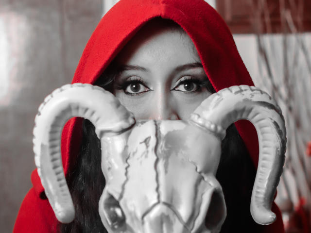 KendraSimons wearing a red hood, eyes staring forward, with a gray, horned skull mask covering her nose and mouth. Her dark, wavy hair is visible under the hood.