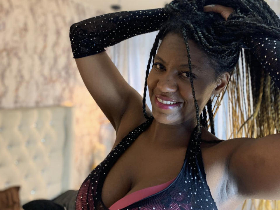 CanelaVelvet with dark skin, long braided hair, smiling, wearing a black, polka-dot, deep-cut top showing her tits, arms raised, in a bright room.