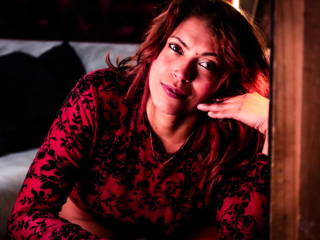 VioletaSexiest with red hair, wearing a red floral top, resting her head on her hand, looking to the side with a subtle smile, glowing in soft, warm lighting.