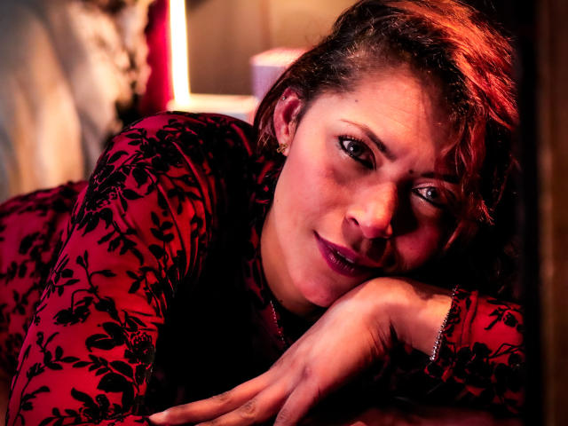 VioletaSexiest with wavy brown hair, red floral dress, and purple lipstick, leans on her hand, looking seductively at the camera with a slight smile. Her ass is partially visible.