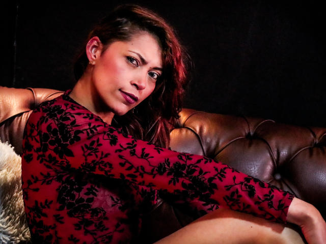 VioletaSexiest, with red floral dress, sits on brown leather couch, her tits slightly visible, dark red lipstick, wavy brown hair, black background, sexy and confident.