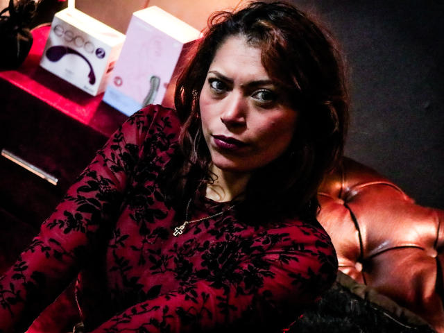 VioletaSexiest has dark brown hair, wearing a red lace top, looking serious with purple lipstick, sitting on a brown leather couch, holding a necklace, in a dimly lit room.