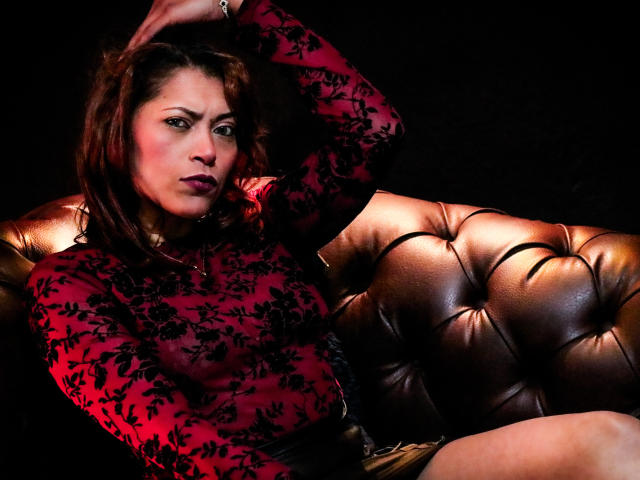 VioletaSexiest with red floral lace top, brown hair, and dark makeup, sits on a brown leather tufted couch, hand in hair, looking seductive, dark background.