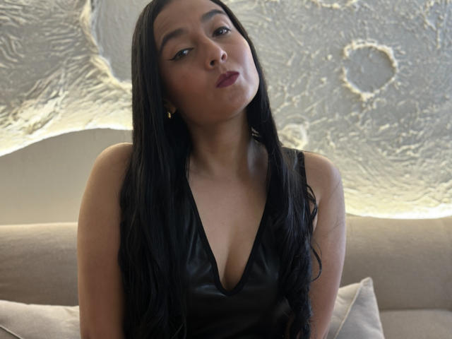 Anthonella Green, met lang zwart haar, draagt een zwarte leren jurk met een diepe V-hals, heeft volle lippen en gouden oorbellen, en kijkt iets omhoog. Haar borsten zijn klein en ze zit op een bank..