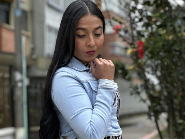 An AntonellaGreen with long black hair stands in an urban setting, looking down with a slight frown. She wears a light blue denim jacket, dark lipstick, and a white wristwatch. Her right hand touches her chin. The background is blurred, showing greenery with red and yellow flowers. The composition focuses on her upper body and facial expression. Her dark hair contrasts with the light blue jacket. The image has a natural, realistic style.