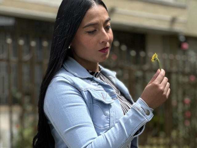An AntonellaGreen with long black hair holds a small yellow flower near her face. She's wearing a light blue denim jacket over a striped shirt. Her dark red lipstick contrasts with her light brown skin. The denim jacket has visible stitching and a small pocket. She's looking down at the flower with a calm expression. The background is blurred, showing a metal fence and some pink flowers. The image is sharp with a shallow depth of field, focusing on AntonellaGreen.