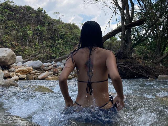 NicoleFoxe stands in a rocky stream, back to the camera, in a black string bikini, with long black hair, tattoo on her back, and wet skin.
