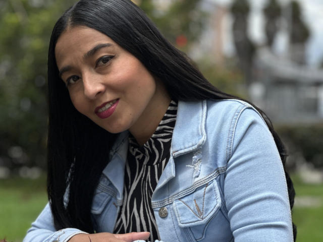 An AntonellaGreen with long black hair, slight smile, and pink lipstick. She wears a blue denim jacket with a small decorative patch and a black-and-white striped shirt. Her light brown skin contrasts with the denim. Blurry green background. She looks relaxed, with hands partially visible. Image has a natural, candid feel. Slight shadow on her face. Casual, everyday style.