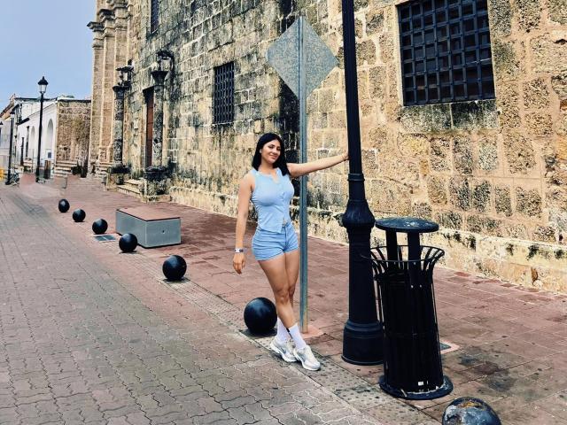 MaddieDemie stands in a sunlit stone alley, wearing a tight blue tank top and shorts, white socks, and white sneakers, leaning on a black lamppost, with a sign.