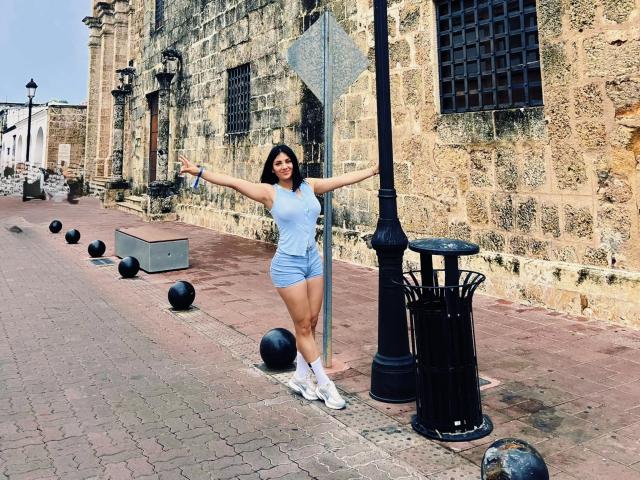 MaddieDemie stands with arms out, wearing a light blue tank top and shorts, white socks, and sneakers, against a stone wall with black bollards and a lamppost.