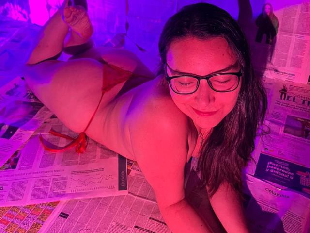 LeilaMoons, nude, with red thong, lying on newspaper, purple and pink lights, glasses, long brown hair, smiling, butt up, legs crossed, soft light, slightly chubby, cute.