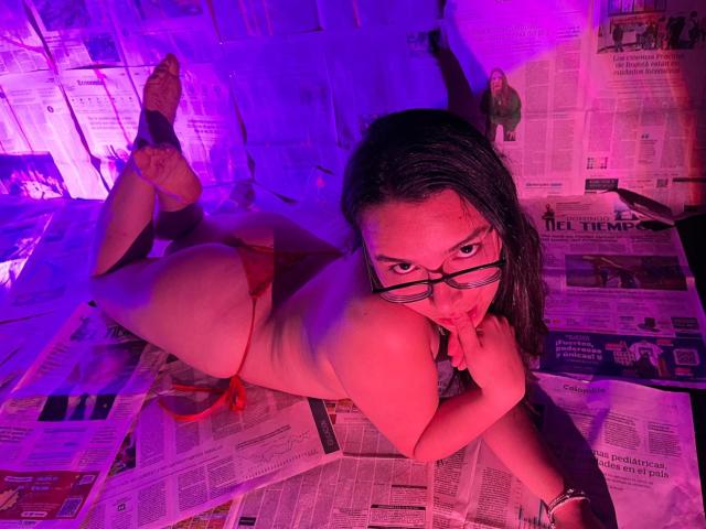 LeilaMoons, naked, lying on newspaper, red thong, glasses, black hair, purple and pink lighting, finger in mouth, butt up, socks, looking at camera, slightly biting finger.