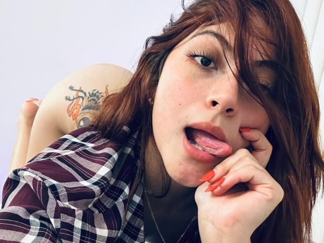 SpencerLe, con i capelli castani, una camicia a quadri, unghie rosse, la lingua fuori, che guarda la fotocamera, mostrando un tatuaggio sulla guancia destra del sedere. Il suo viso è vicino alla fotocamera..