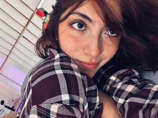 SpencerLe tiene ojos azules, piel clara y cabello castaño, lleva una camisa de cuadros, mira hacia arriba con una ligera sonrisa, mostrando sus labios fruncidos. Sus pechos son parcialmente visibles..