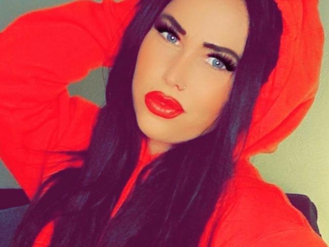 NastyBitch81-hot with red lipstick, blue eyes, and dark hair, wearing a red hoodie, with a nose ring, and a serious expression, holding her hair back.