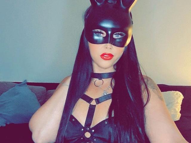 NastyBitch81-hot with long black hair, wearing a black leather mask with ears, red lipstick, black harness, and choker, sitting on a couch with blue and gray pillows.