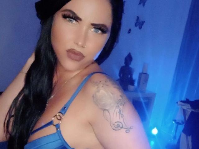 NastyBitch81-hot with blue eyes, dark hair, and a nose ring, wearing a blue strappy top, showing a tattoo on her right shoulder, in a blue-lit room.