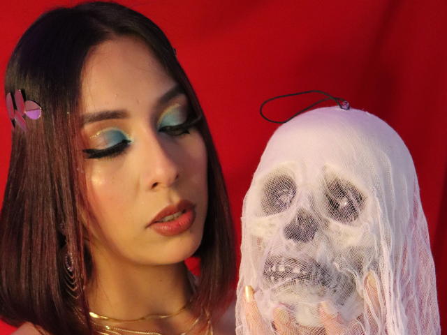 MoollyEvanns met blauwe oogschaduw en lange wimpers, die een wit schedelmasker vasthoudt met een zwarte koord, voor een rode achtergrond. MoollyEvanns heeft bruin haar en roze lippenstift..