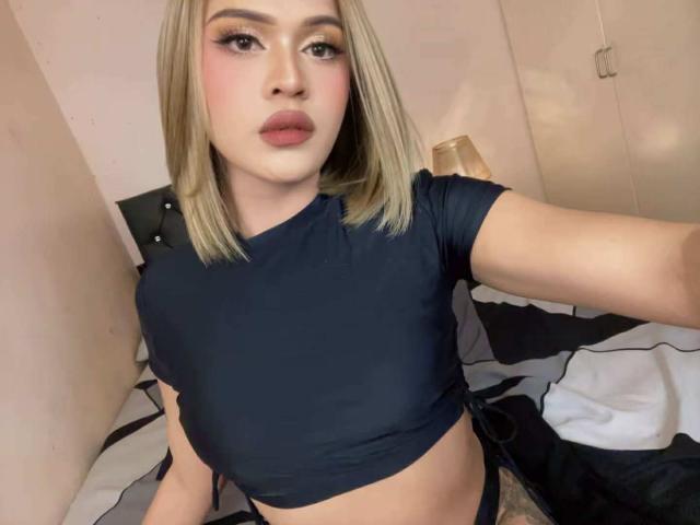 TsQueenxMia with blonde bob, wearing a black crop top, showing her tits, with pink makeup and pouty lips, taking a selfie in a bedroom.
