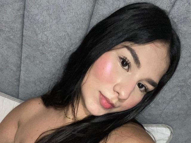 DahliaRosse has long black hair, pink blushed cheeks, and brown eyes. She's looking at the camera with a slight smile, wearing a gold necklace, and has bare shoulders.