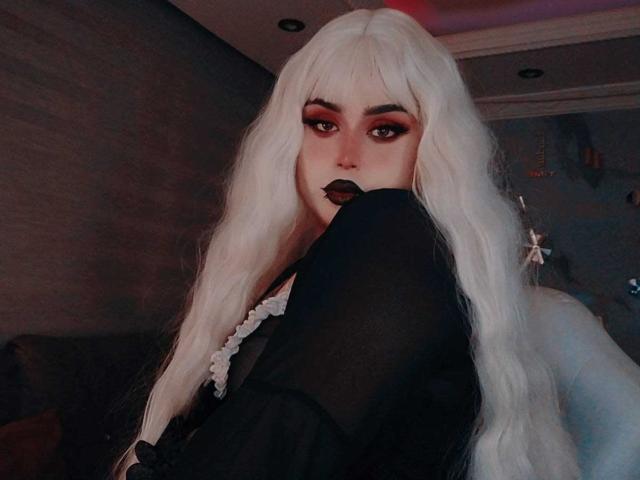 MadameRaven with long white hair, dark makeup, and black lipstick, showing off her black thigh in a dark room, wearing a black top with white lace.