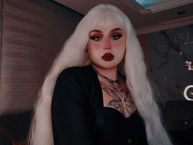 MadameRaven with long white hair, dark red lipstick, and black top, showing tattoos on her chest, looking directly at the camera. Her eyes are heavily made up.